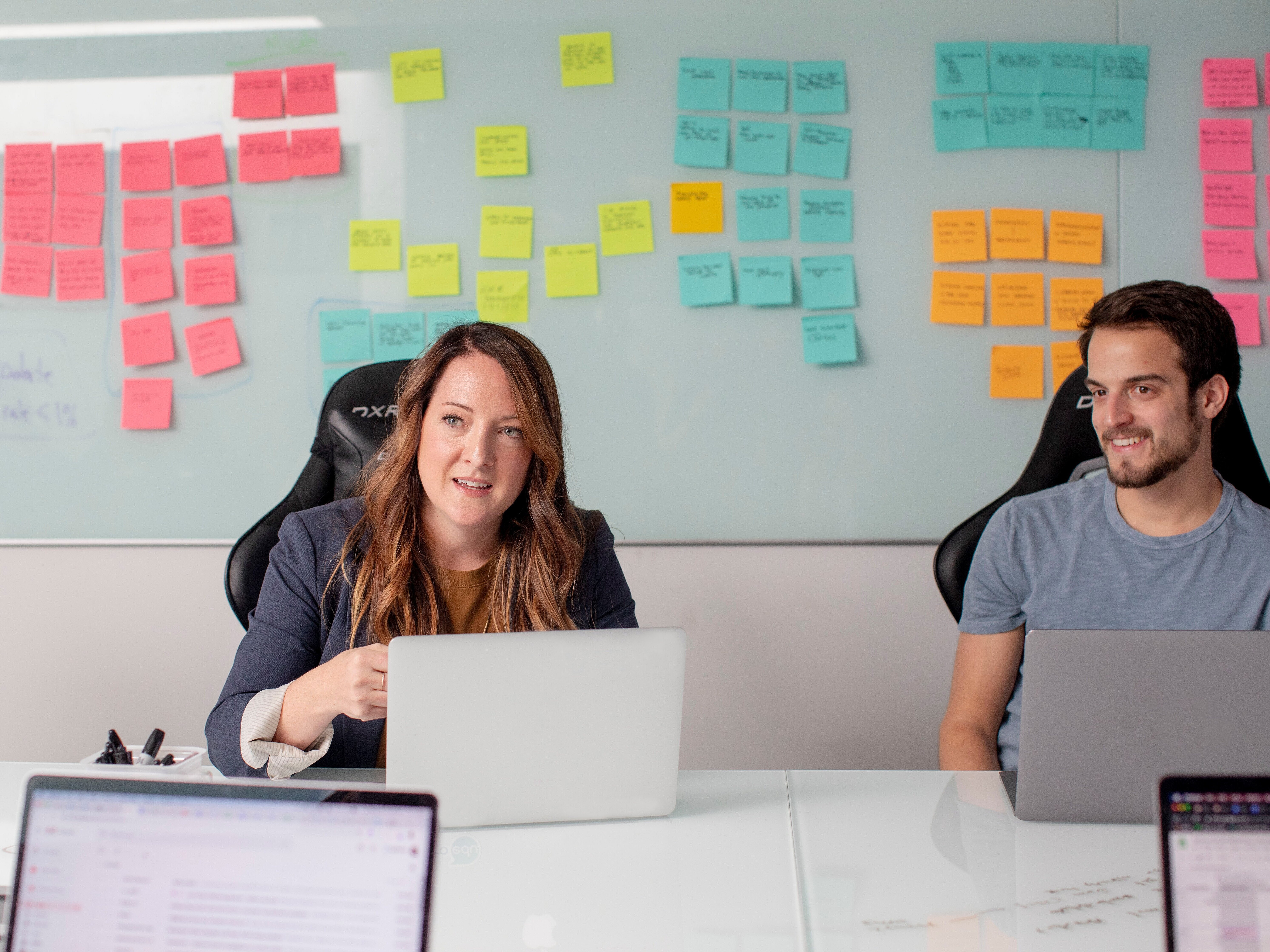 Eine Frau und ein Mann mit Laptops an einem Meetingtisch, im Hintergrund ein Whiteboard mit bunten Post-its